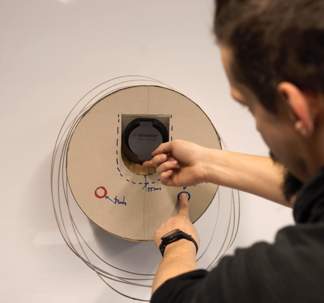 zdjęcie przedstawia tekturową makietę okrągłej ładowarki do samochodu elektrycznego i fragment sylwetki projektanta sprawdzającego jej funkcjonalność